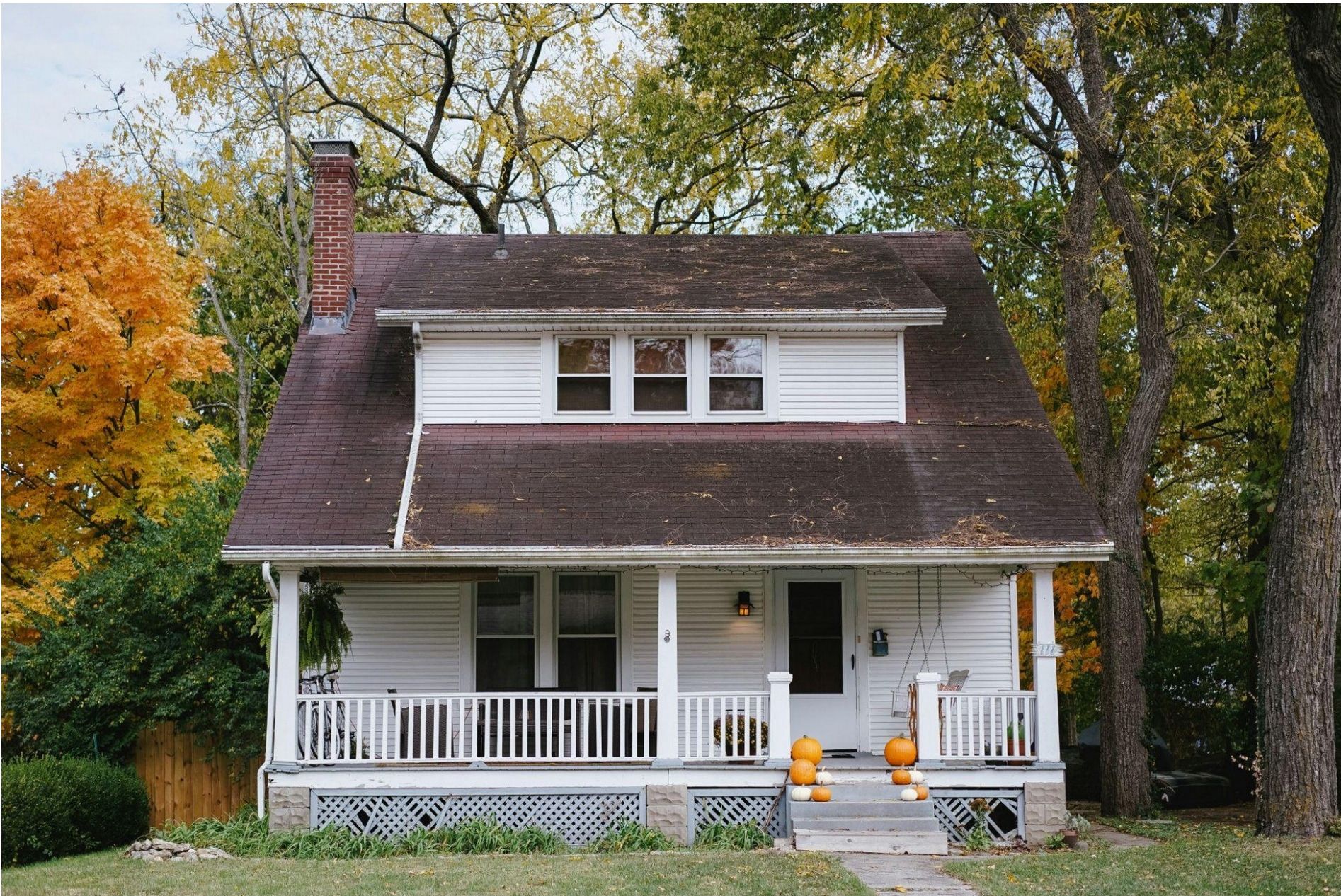 A white house with a brown roof and a front porch surrounded by autumn trees