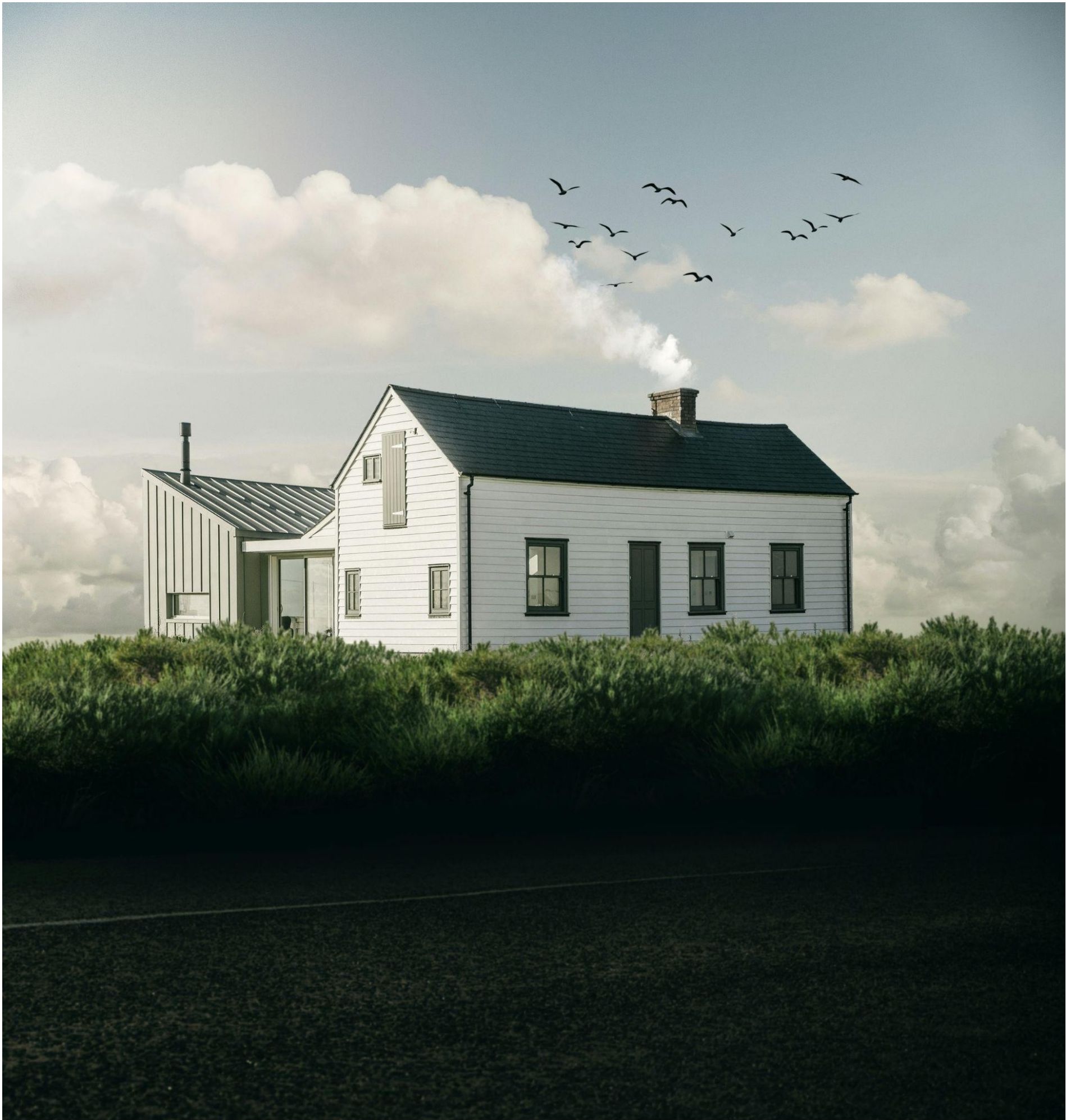A white house with a dark roof, surrounded by green bushes and a field, under a cloudy sky with birds flying.