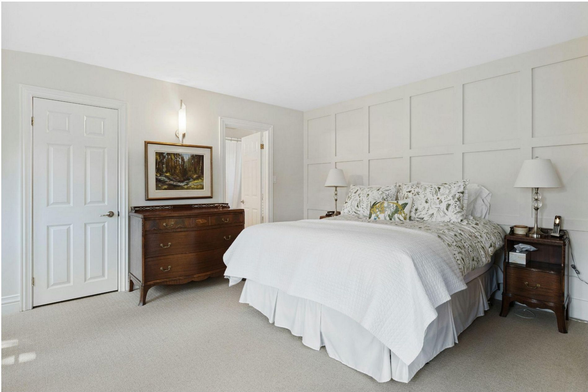 A bright, minimalist bedroom featuring a large bed with white linens, a dark wood dresser, and paneled walls.