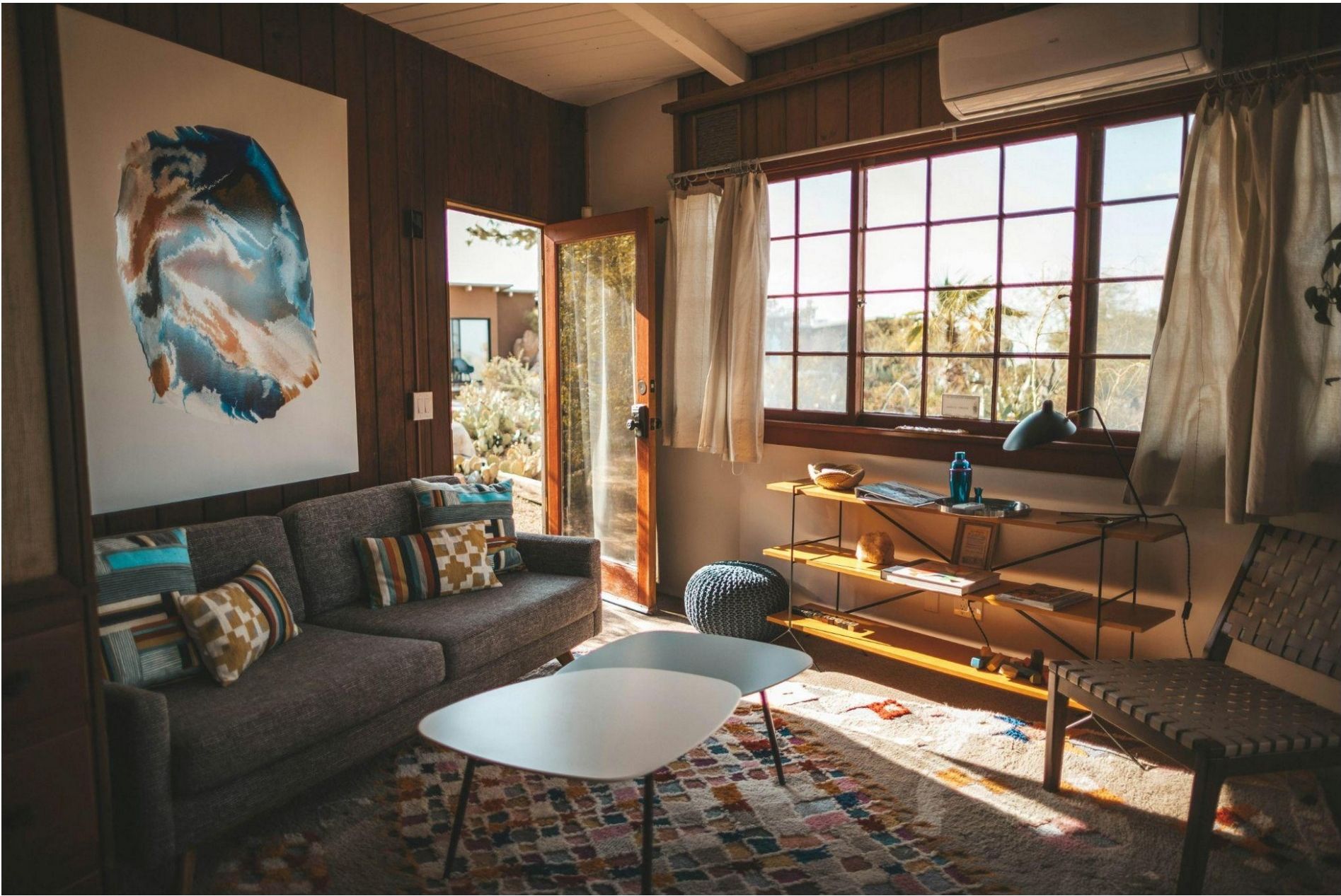 A cozy living room with a large window, wooden walls, a grey sofa with colorful pillows, a white coffee table, and a desk by the window